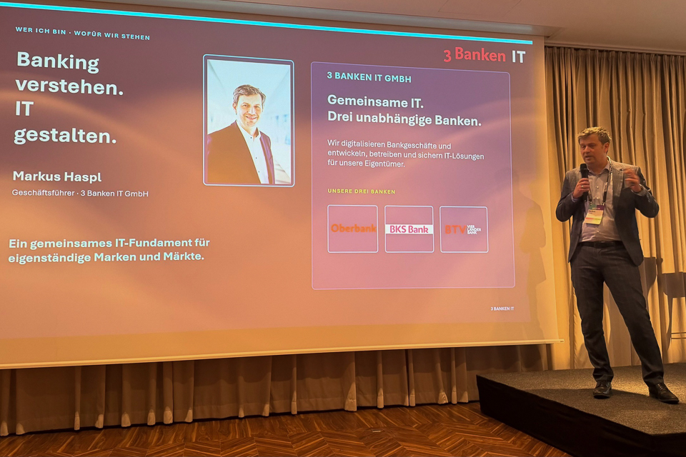 Geschäftsführer Markus Haspl steht auf einer Bühne und hält ein Mikrofon, während er vor einer großen Bildschirmpräsentation spricht, die auch das Logo von „3 Banken IT GmbH“ zeigt.